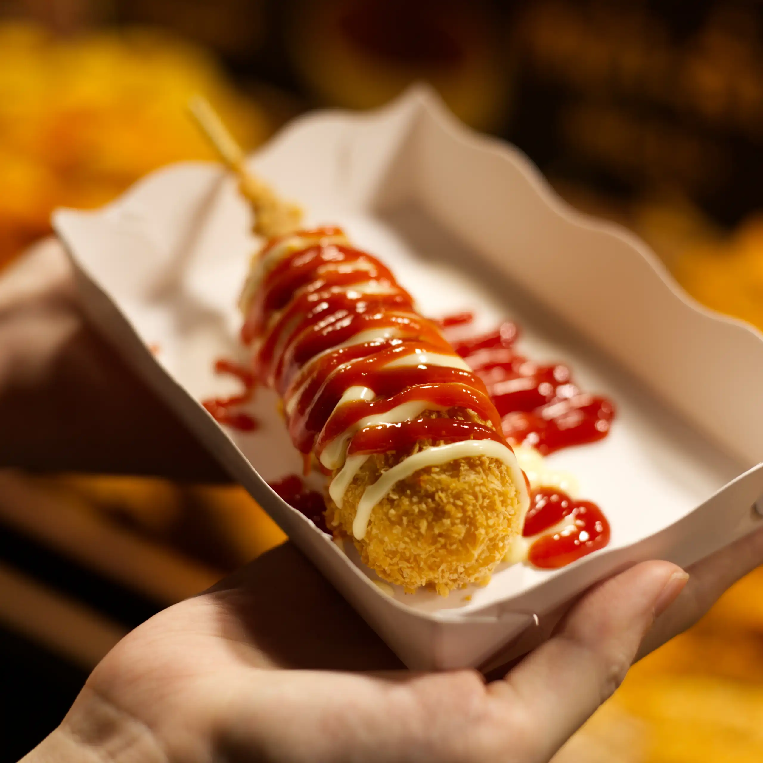Une personne tient dans ses mains une barquette remplie d'un corn dog.
