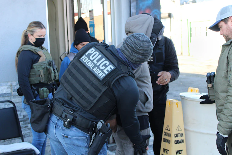 Armed man with jacket that says 'Police Federal Agent' pats down person in sweatshirt and jeans as other people stand watching