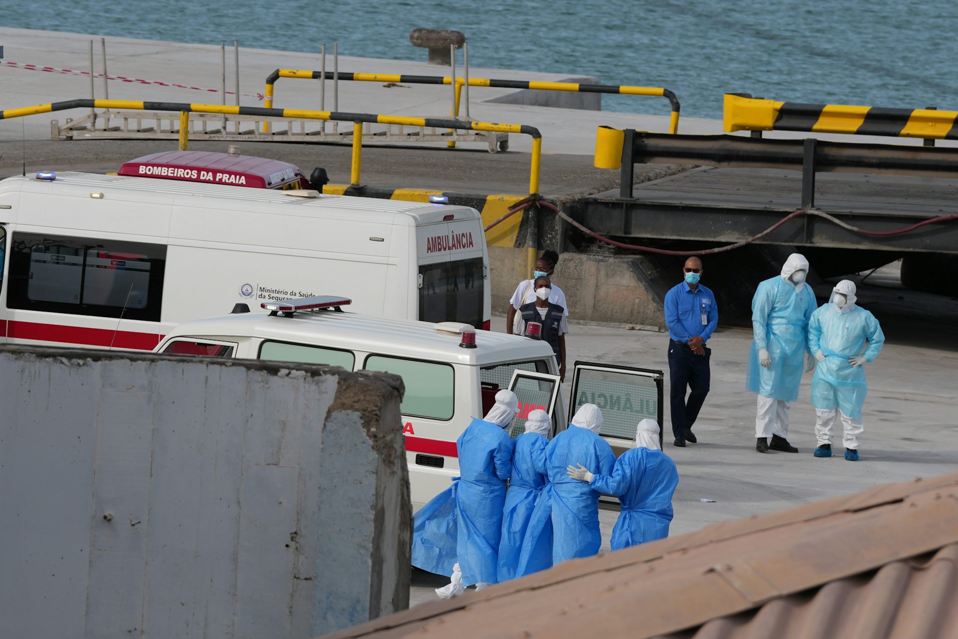 Medical staff in blue gowns on the quay of a seaport, with emergency vehicles around them
