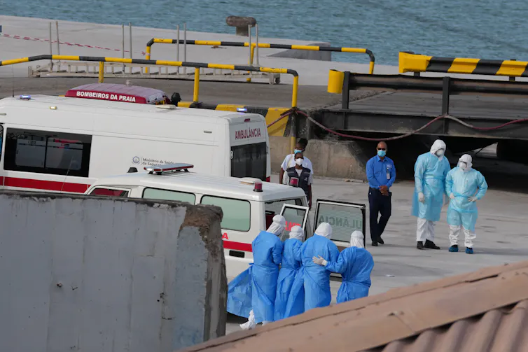 Medical staff in blue gowns on the quay of a seaport, with emergency vehicles around them