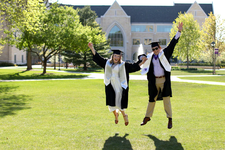 Two people seen jumping on a lawn in graduate caps and gowns.