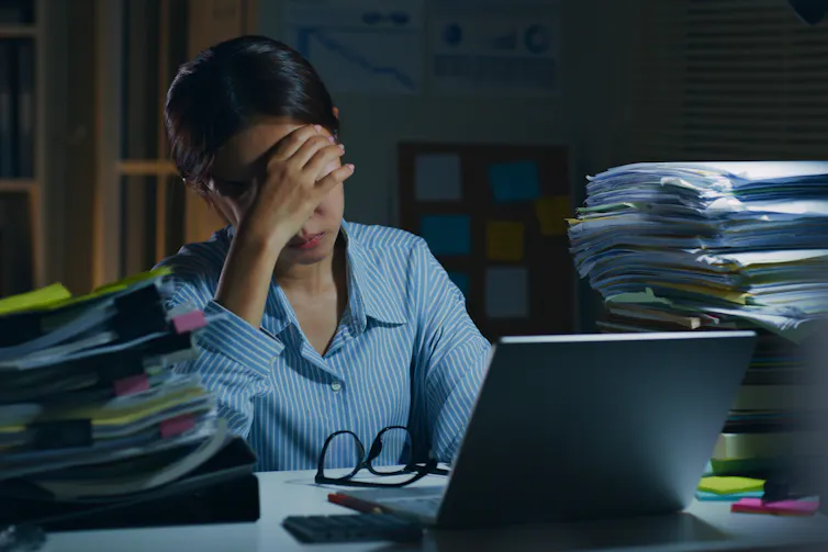 Une femme appuie son front contre sa main, assise à un bureau encombré de paperasse