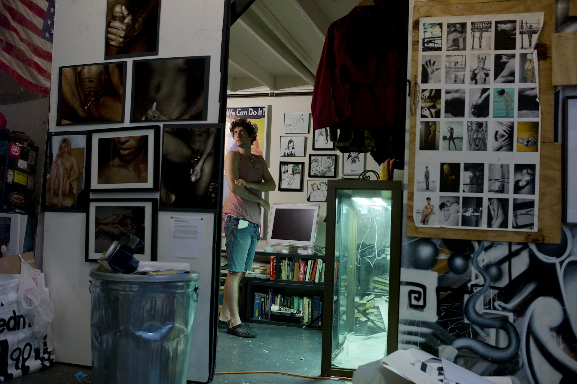 A young man folding his arms is visible through a mirror, which shows a room with walls filled with photographs and other imagery.