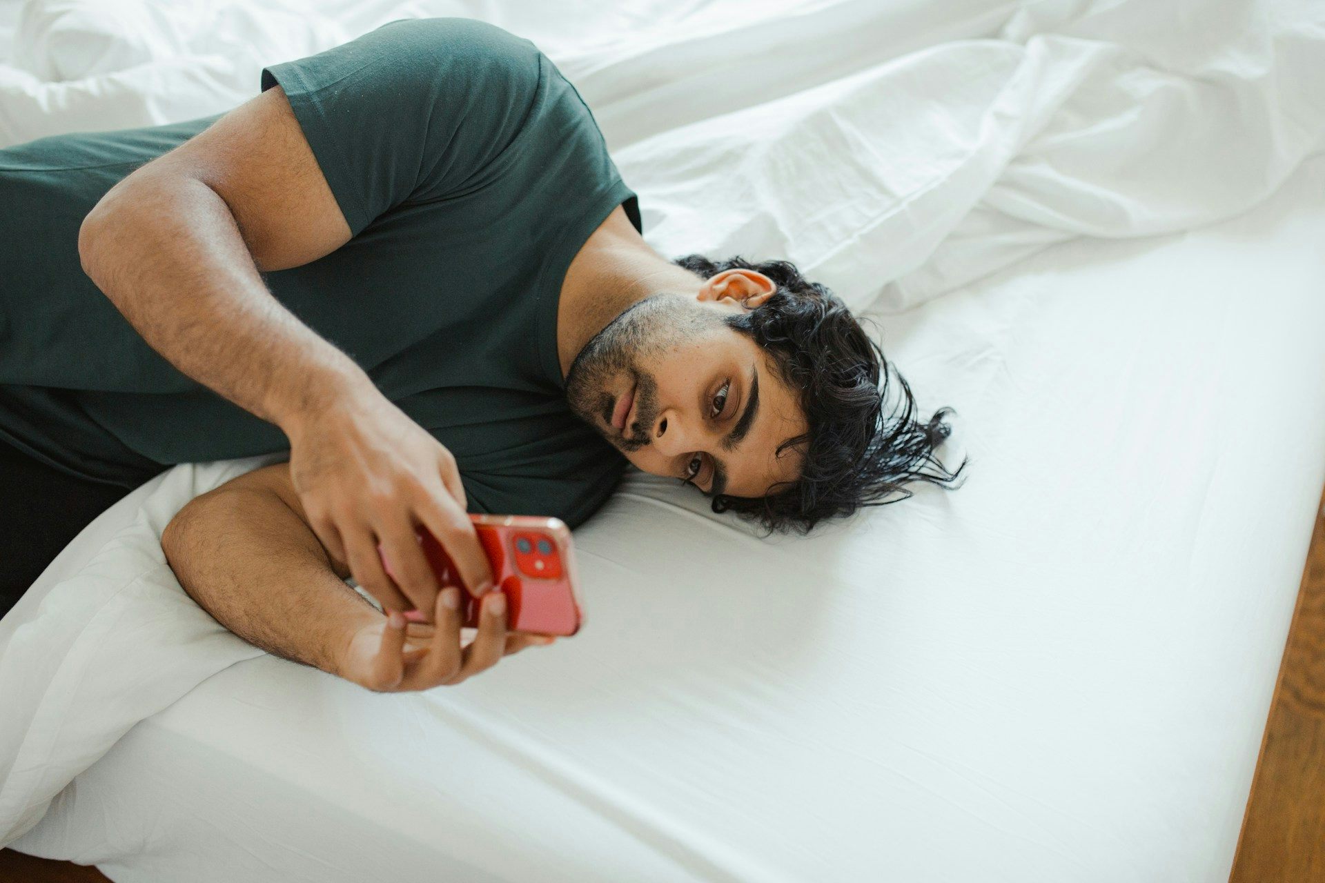 Un hombre yace acurrucado en una cama blanca usando el teléfono.