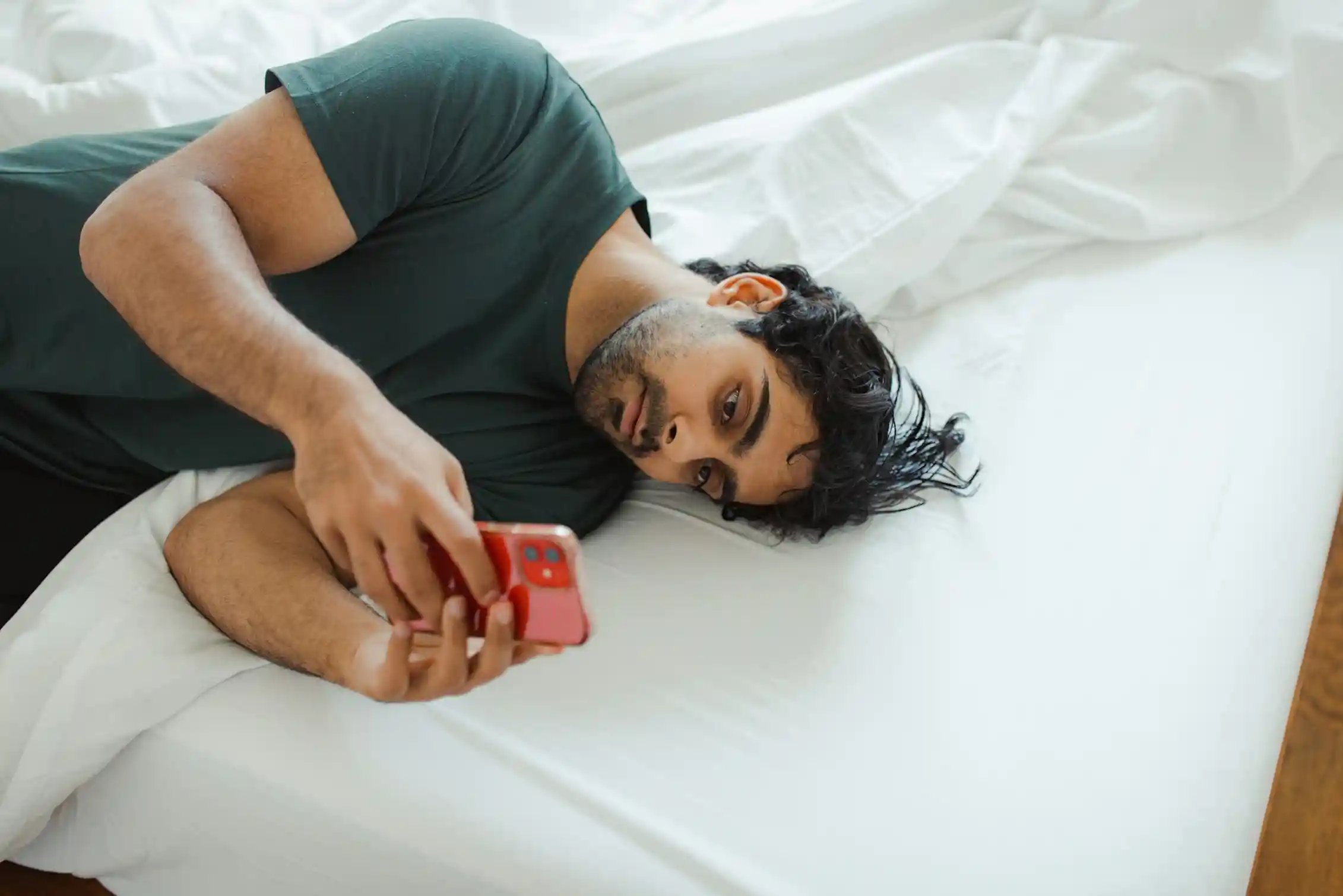 Man lies curled on a white bed using his phone.