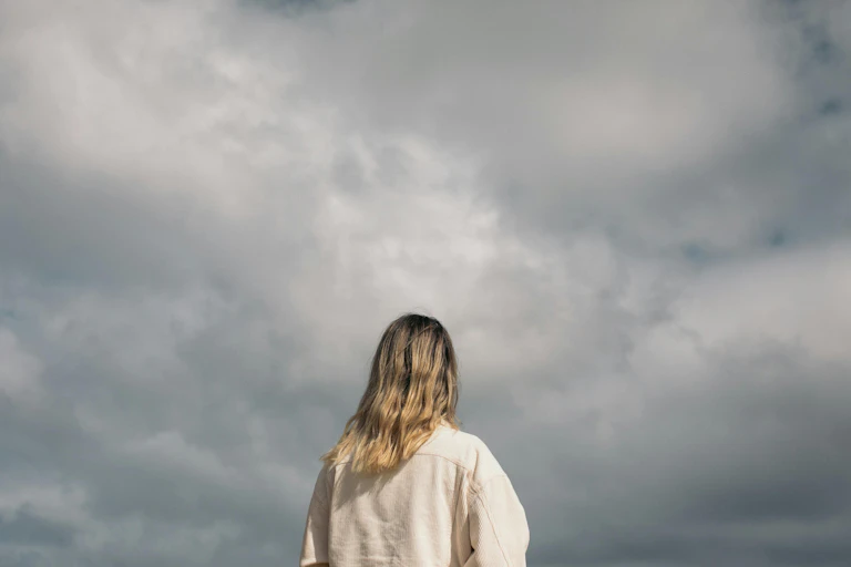 A young person faces a cloudy sky. Their back is to the camera.
