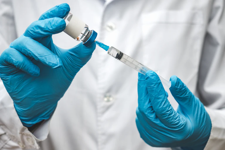 A doctor or nurse wearing blue gloves fills a syringe from a vial of clear liquid.