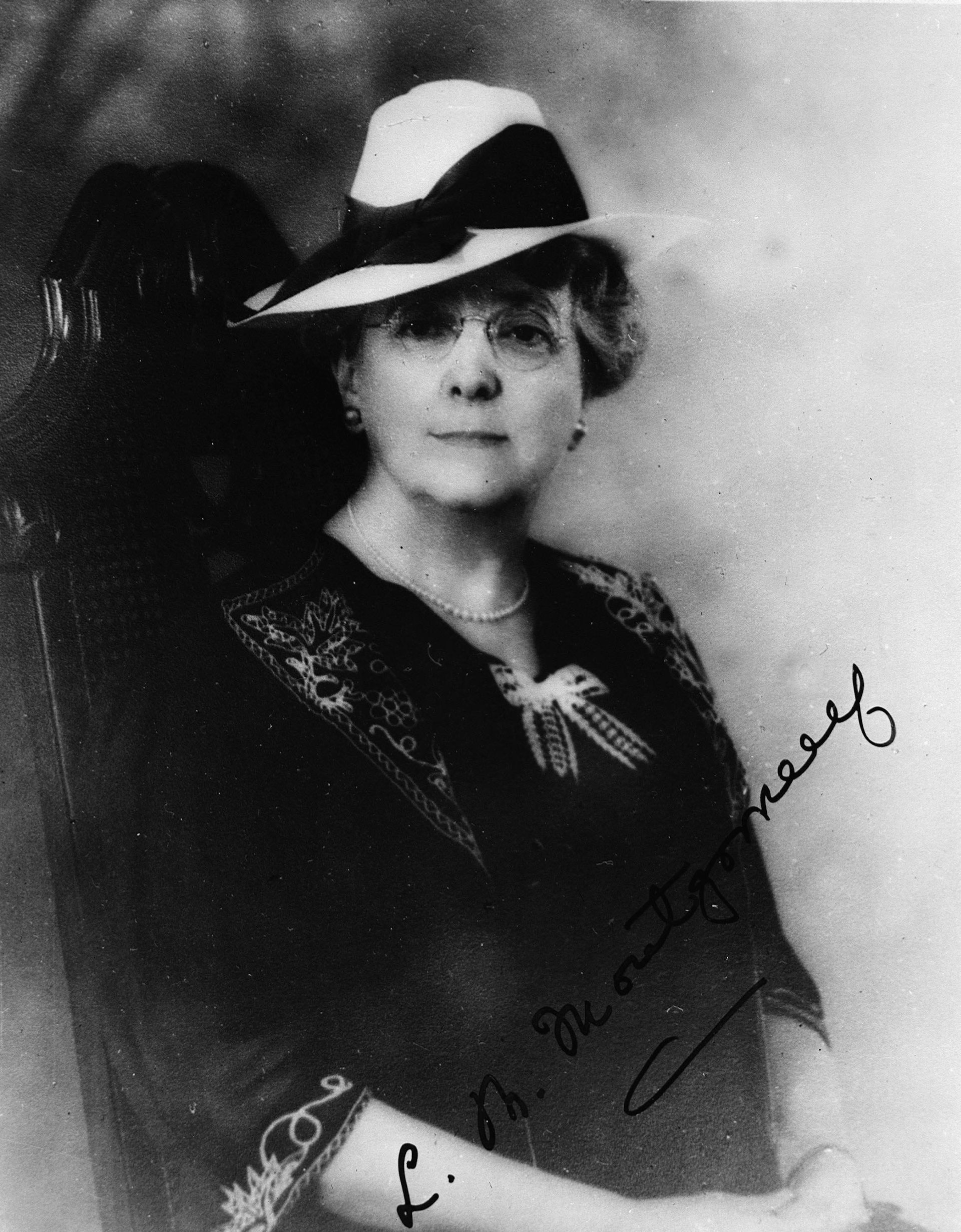 Fotografía en blanco y negro de una mujer con sombrero, gafas redondas y vestido de principios del siglo XX con escote en pico, lazo y manga 3/4 con bordado de follaje/arabescos.