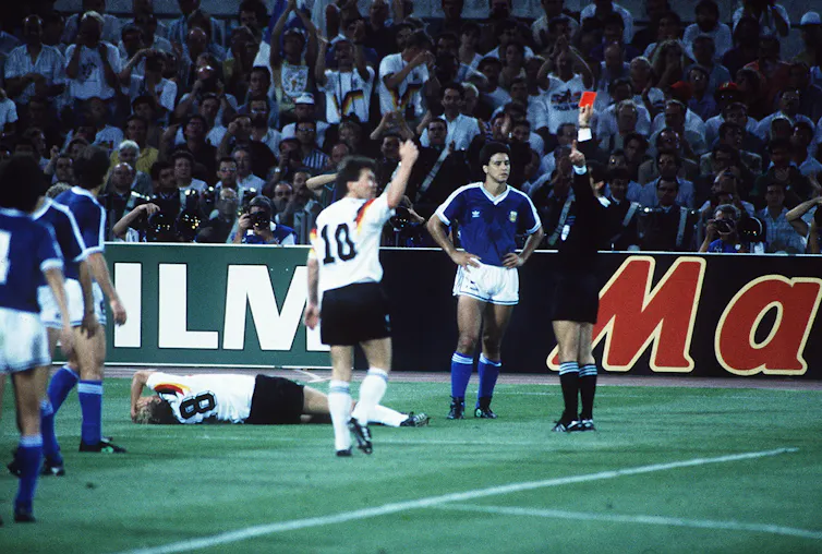 A man in black holds aloft a red card as football players stand around.