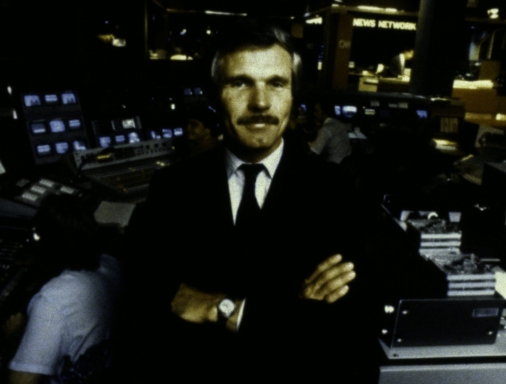 A man stands in a newsroom, arms folded.