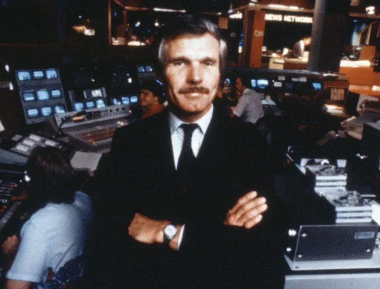 A man stands in a newsroom, arms folded.