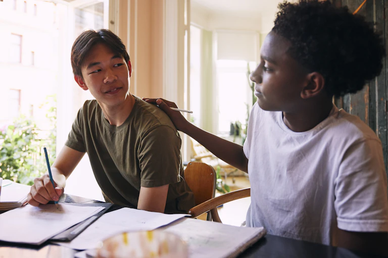 One boy puts his hand on the shoulder of another as they work together at a table