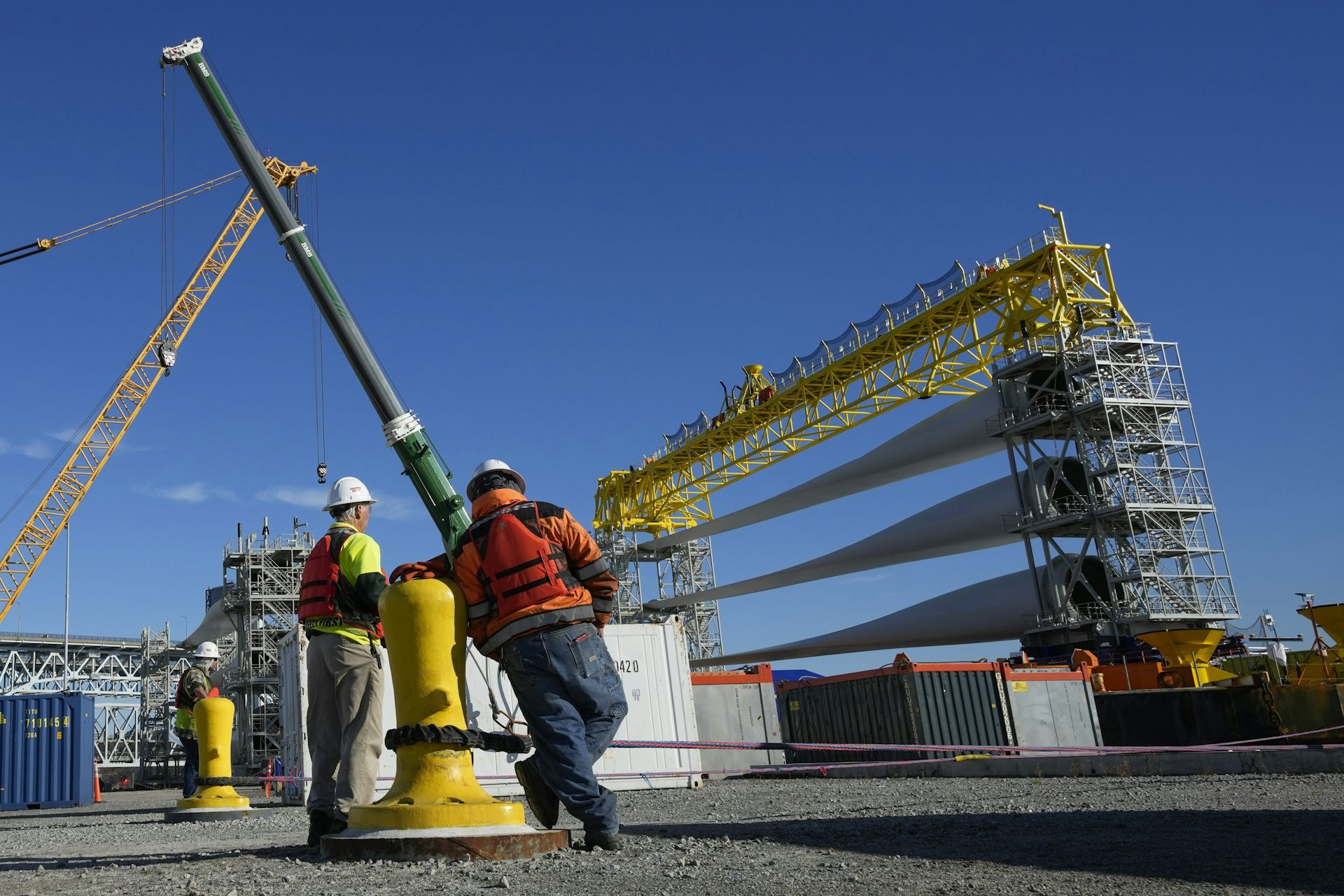 Two workers stand on a dock as wind turbine blades are loaded on a ship with a crane.
