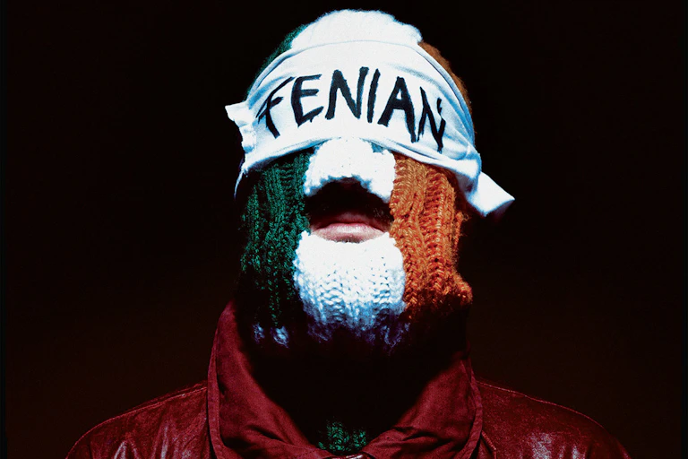A person is shown against a dark background wearing a red jacket and a knitted face covering in the colours of the Irish flag, green, white, and orange. Their eyes are covered by a white blindfold with the word “FENIAN” written across in black letters.
