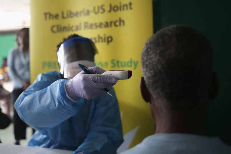 Person in protective clothing aims a device at another person's head