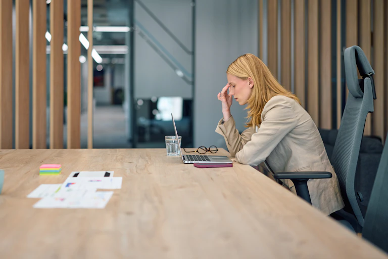 Businesswoman at a table with laptop has closed eyes and holding fingers to her forehead