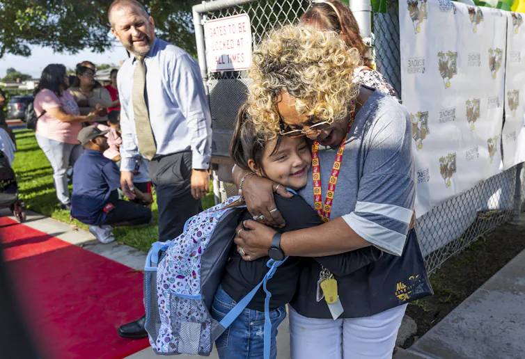 A woman with blonde hair hugs a girl wearing a backpack, and they both smile as a man wearing a tie looks at them and also smiles.