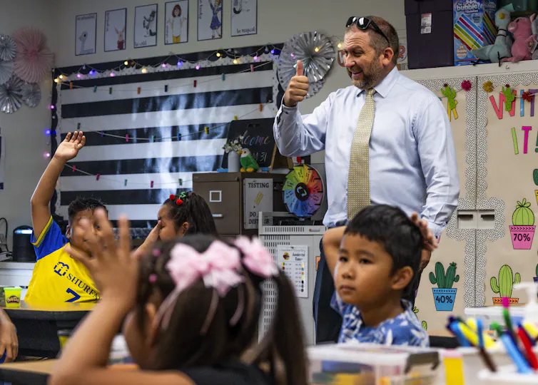A man wears a tie and gives a thumbs up as a group of children seated at desks raise their hands.
