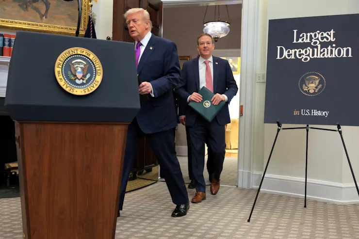 Two men walk toward a podium. One of them, Zeldin, is grinning. The promotional sign reads 'Largest Deregulation in History