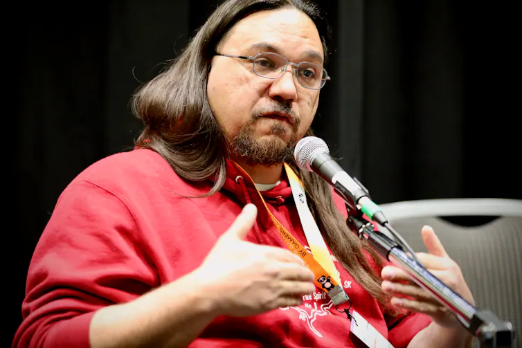 a man with long black hair wearng glasses and a red shirt speakng into a microphone.