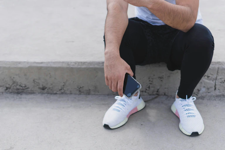 A man in running sneakers and attire holds their phone while sitting down.