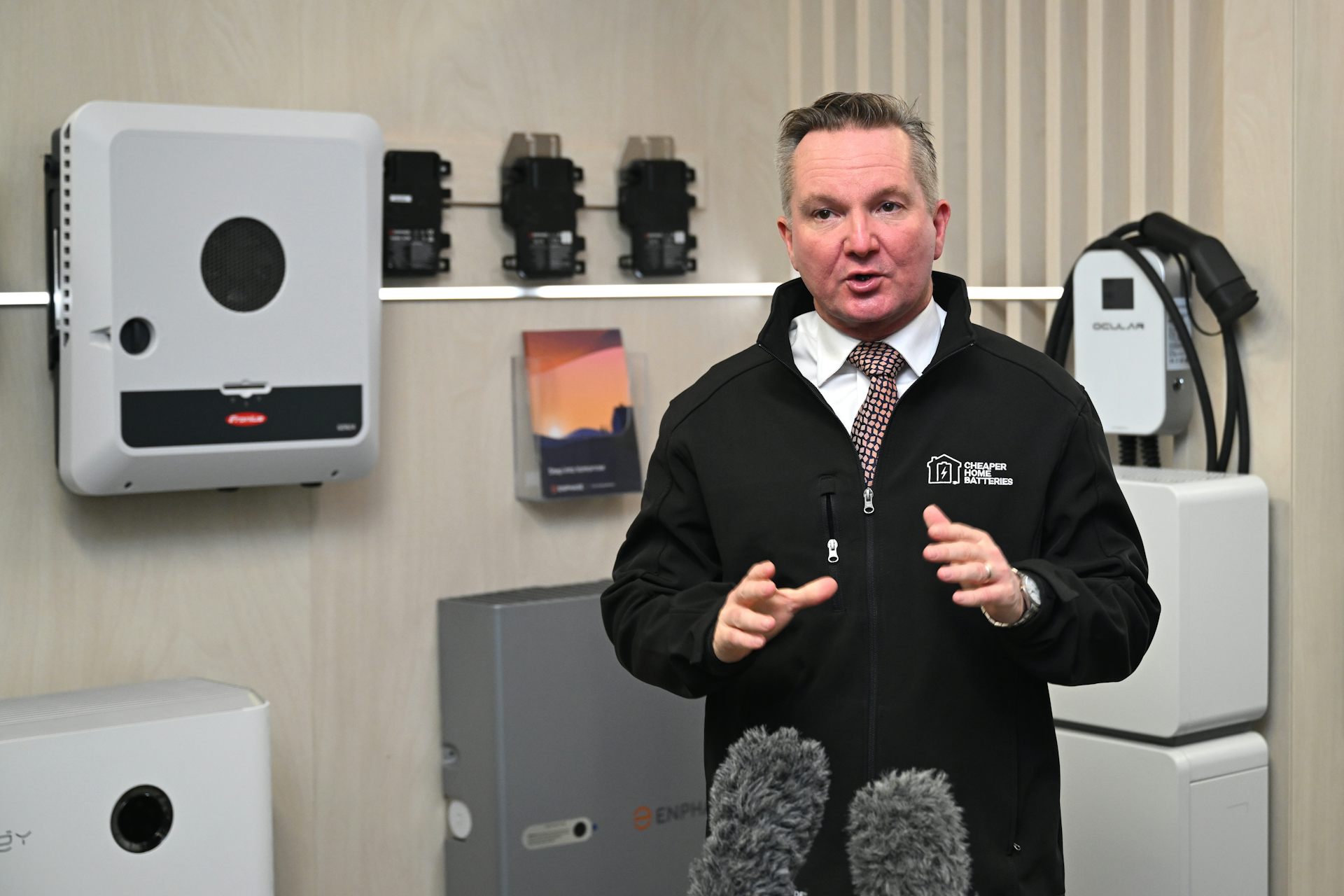 A man in a tie and black jacket speaks to the media in front of a home battery system.