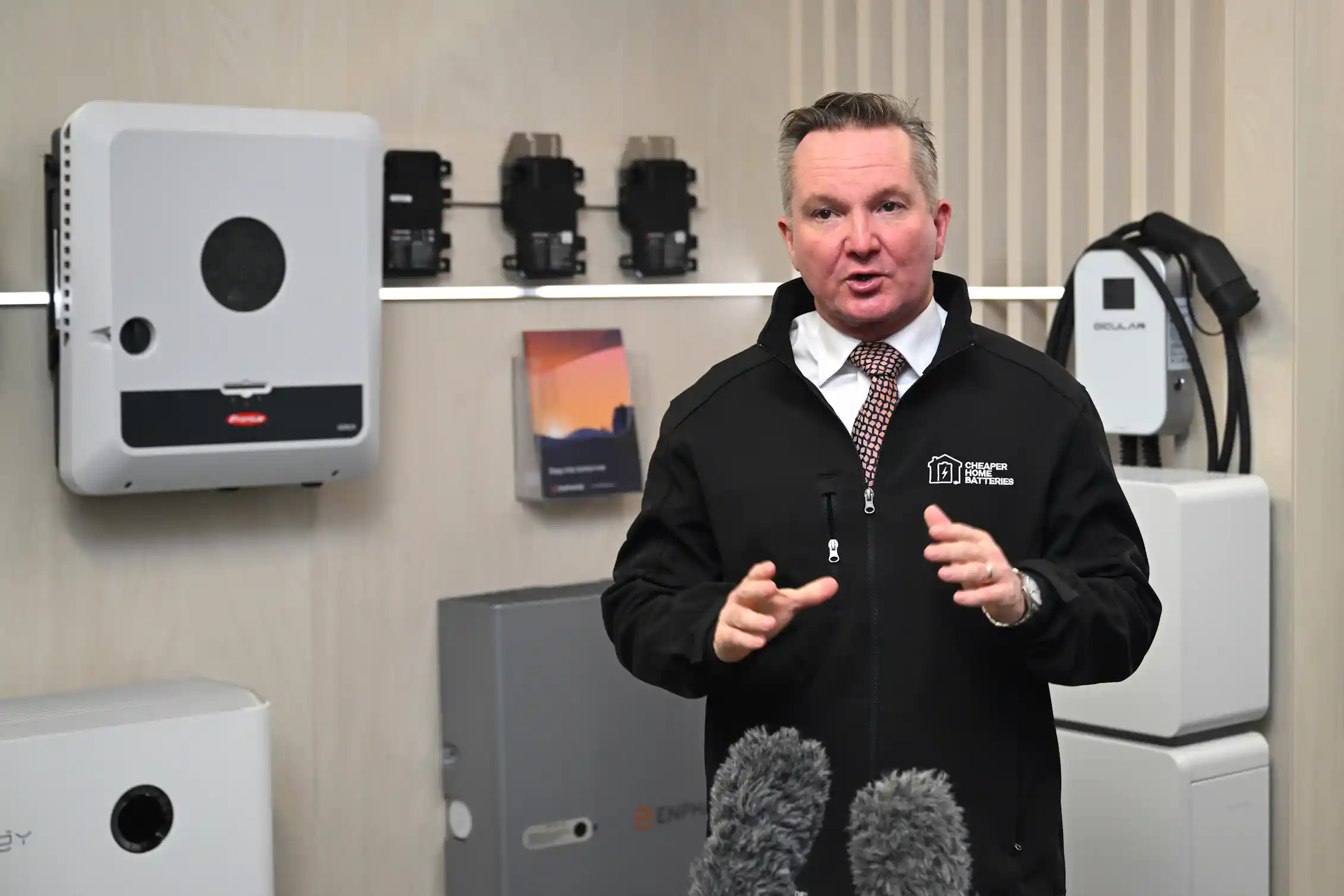 A man in a tie and black jacket speaks to the media in front of a home battery system.