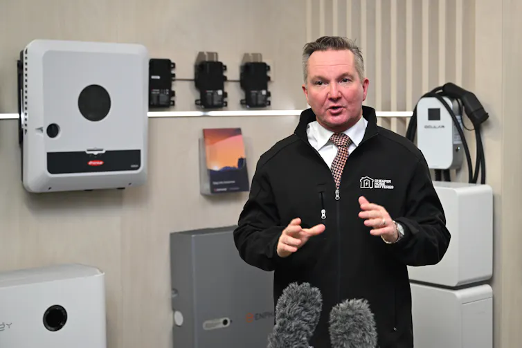 A man in a tie and black jacket speaks to the media in front of a home battery system.