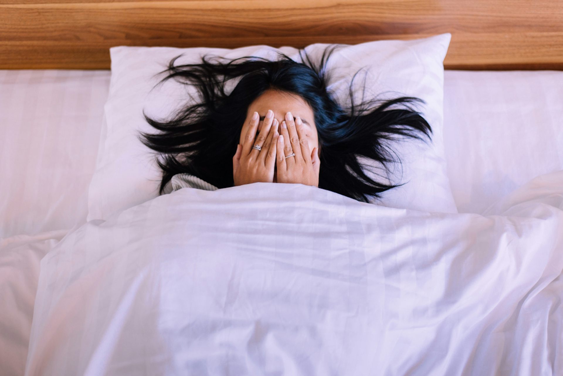 A woman lying in bed, hands over her face, suggesting remembering something bad.