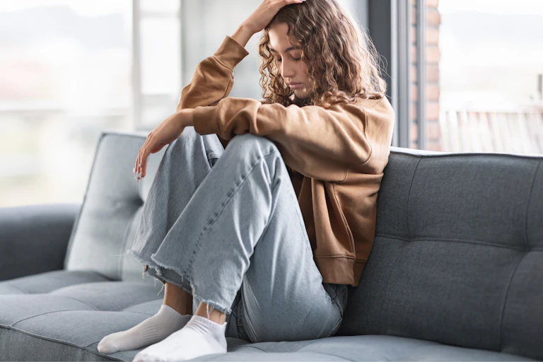 A teenage girl sits on a couch, looking sad or frustrated.