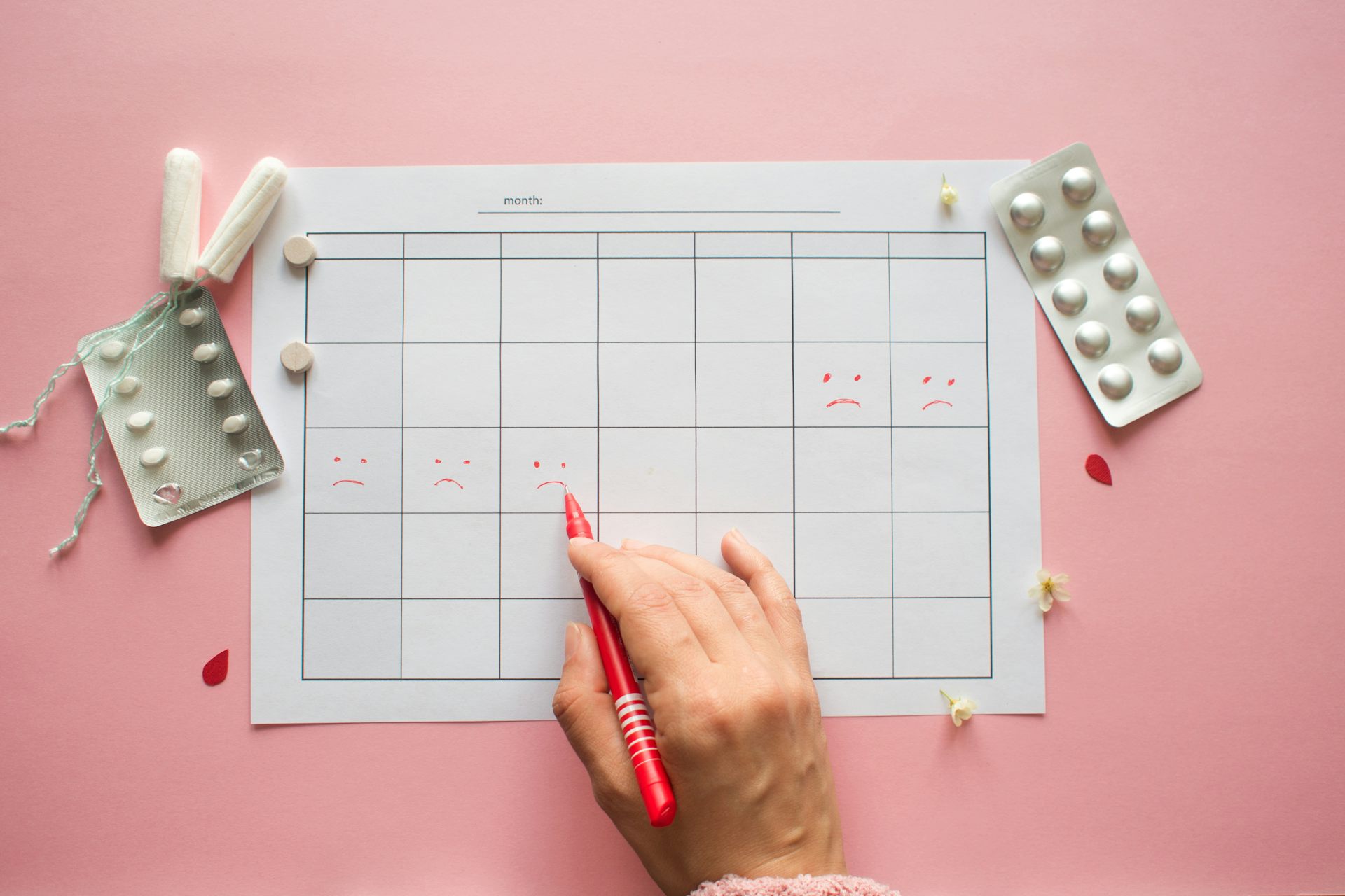 A persons's hand marks a calendar with frowny faces over five-days to depict menstruation.