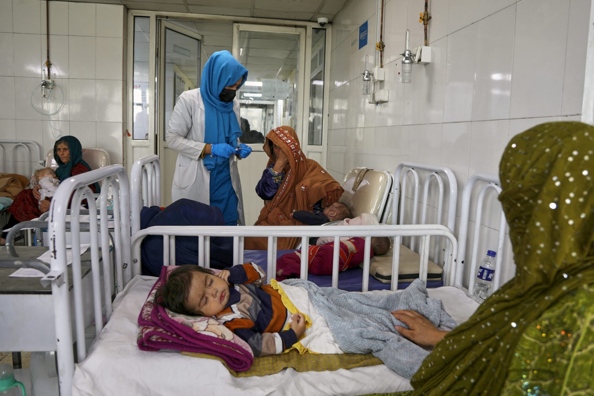 Women in head scarves sit near a hospital bed where a young baby sleeps.