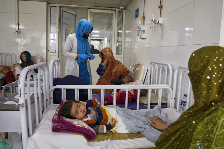 Women in head scarves sit near a hospital bed where a young baby sleeps.