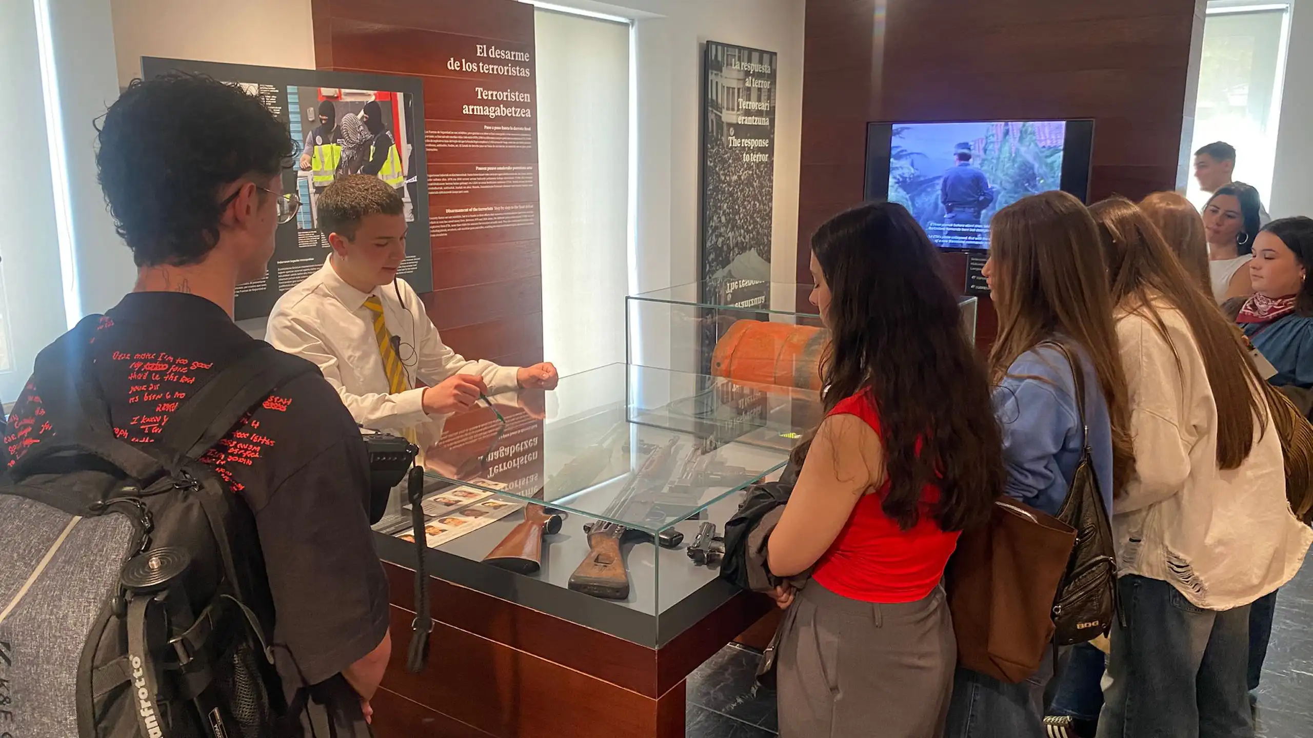 Un grupo de jóvenes entorno de un expositor frente a un docente de un museo.