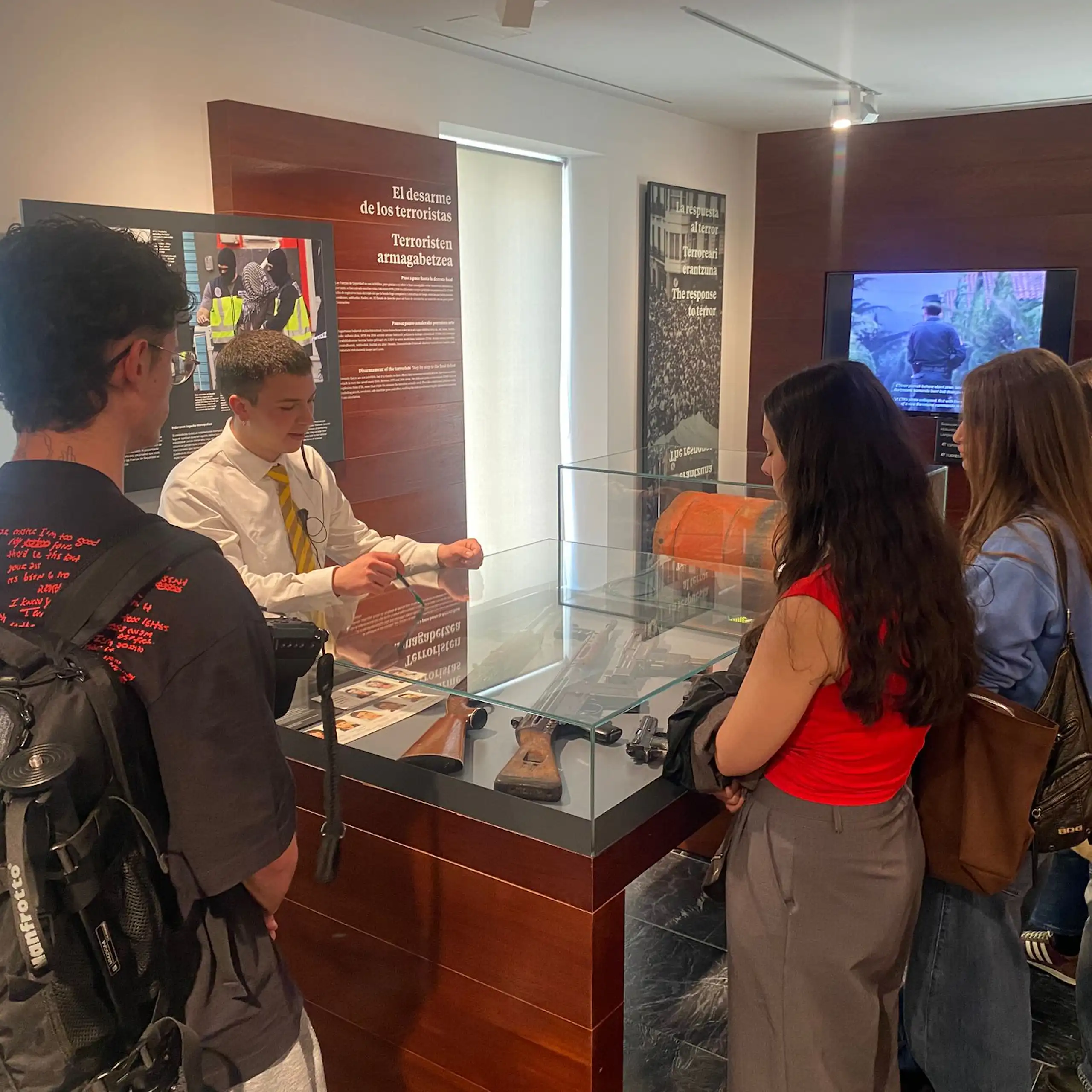 Un grupo de jóvenes entorno de un expositor frente a un docente de un museo.