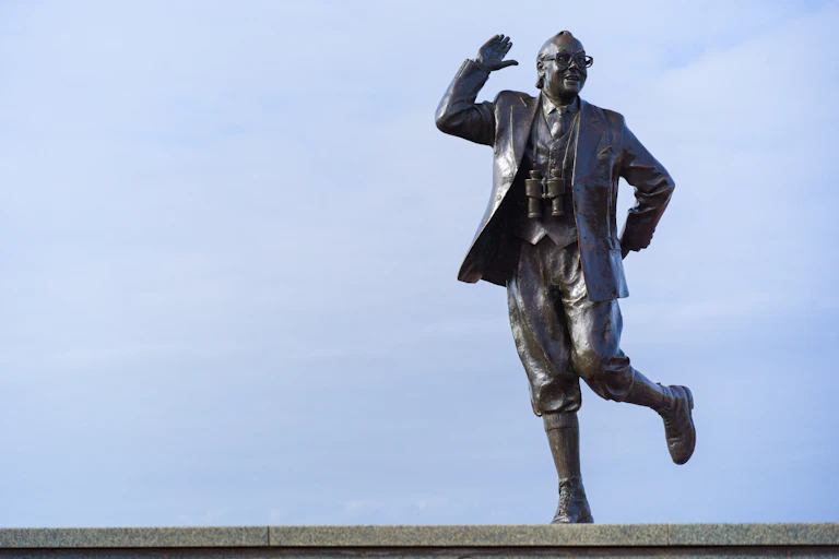 A statue of Eric Morecambe doing the Bring Me Sunshine dance against a blue sky.