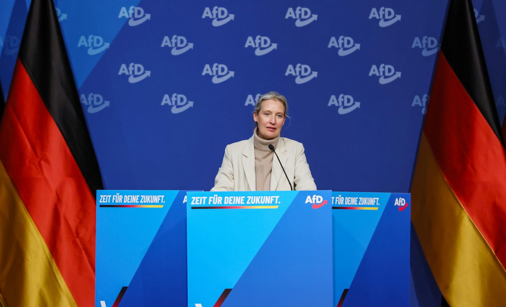 Alice Weidel speaks at an event flanked by German national flags.