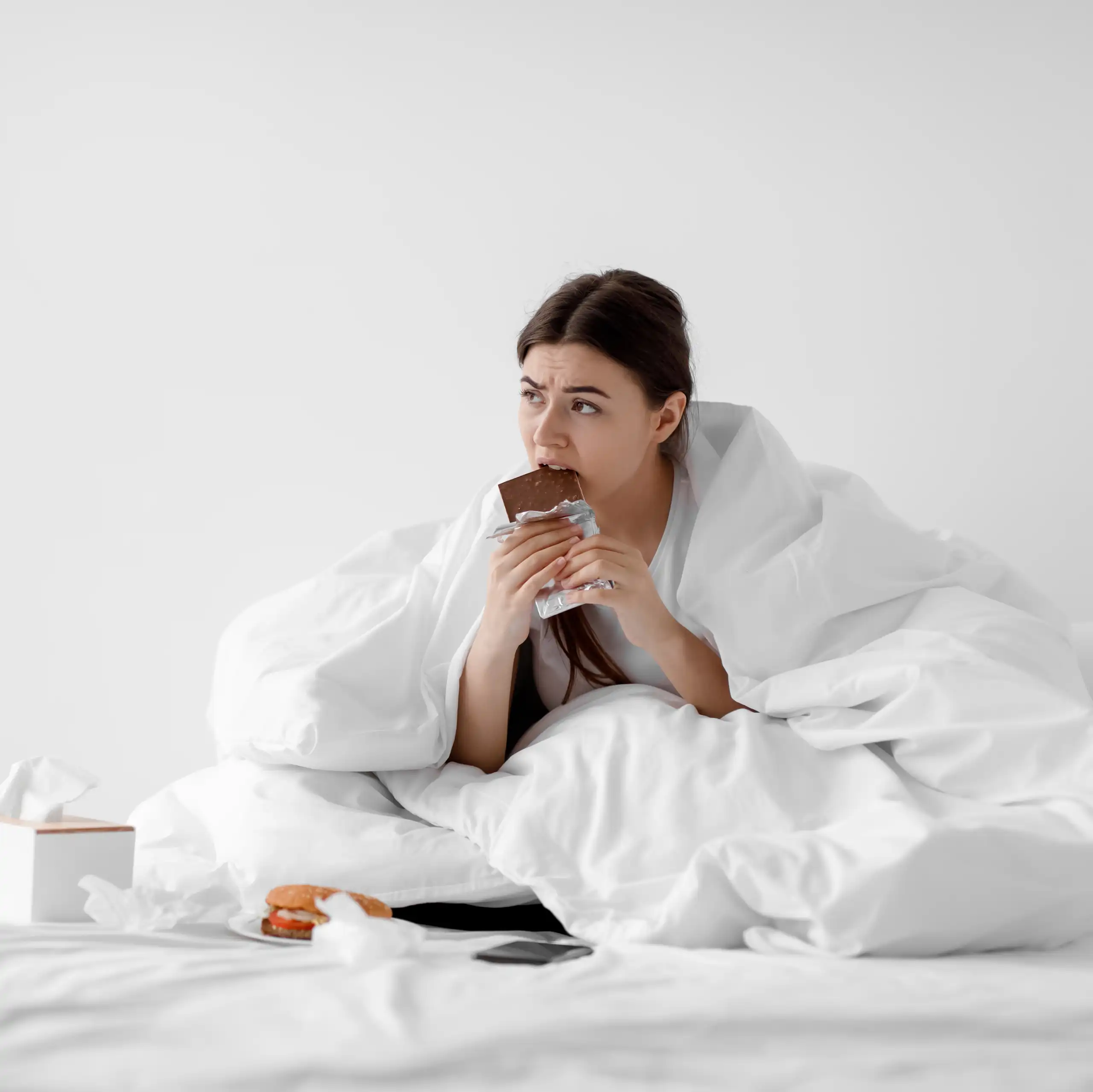 Una xica menja xocolata en el llit tapada amb un llançol.
