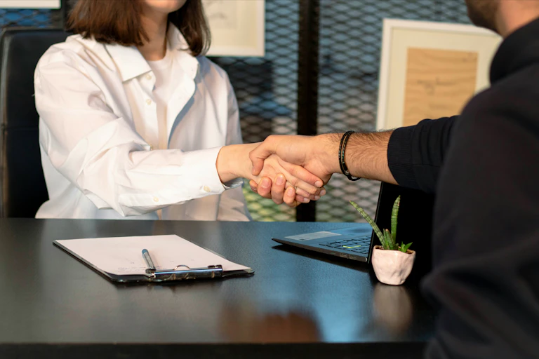 A woman and a man, seen from the neck down, shake hands across a desk