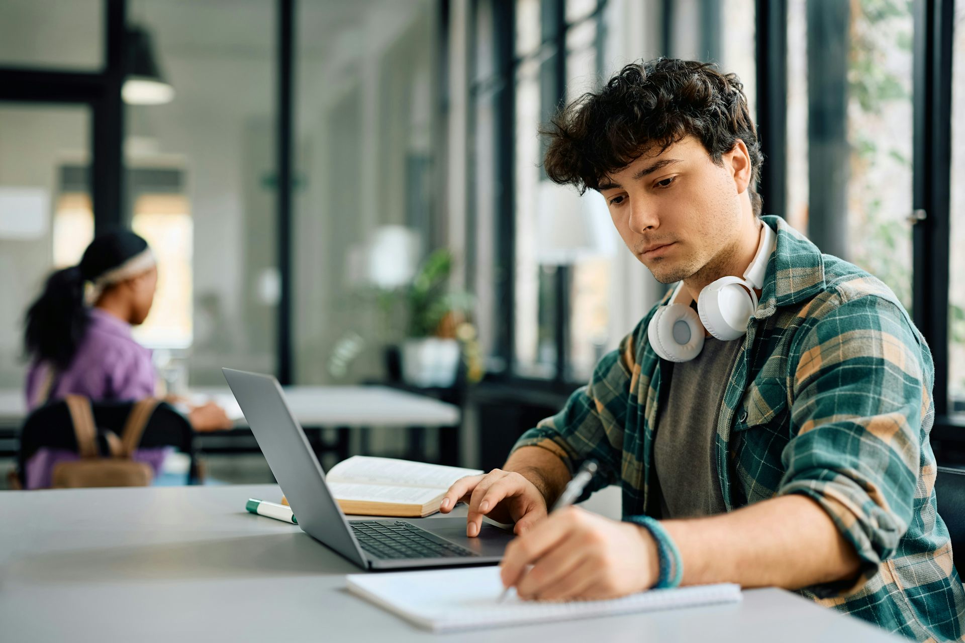 un joven se sienta en una mesa, trabaja en una computadora portátil y escribe en un cuaderno