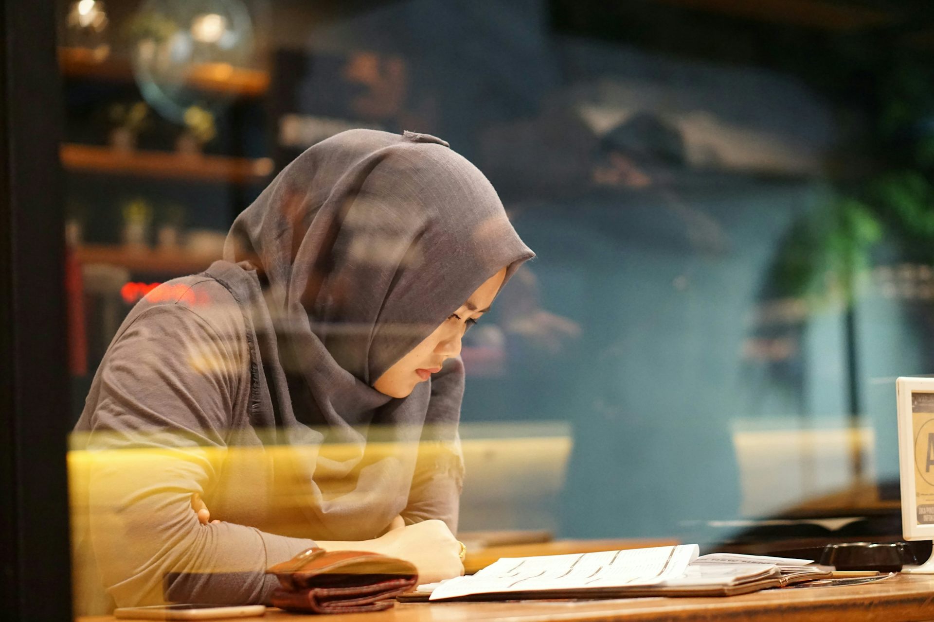 a young woman wearing a headscarf sitting at a table reading a book