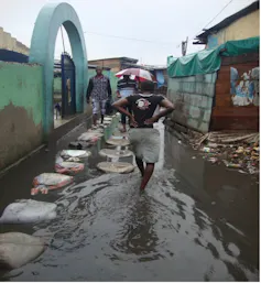 A woman wades through an area between houses where water is above her ankles with a line of sandbags set up for people to step on