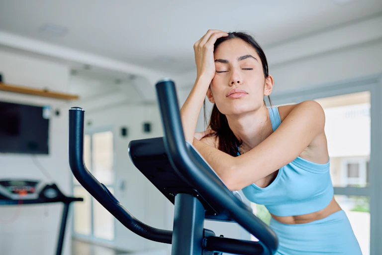 A tired woman closes her eyes and rests while using a stationary bicycle at the gym.