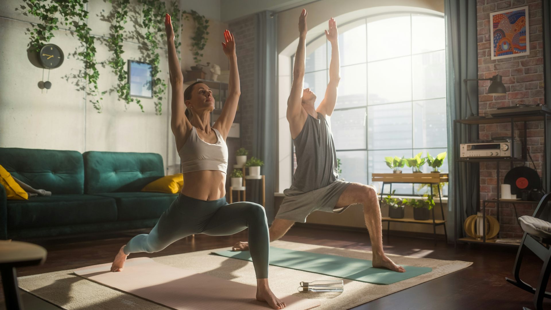 A fit man and woman perform a yoga move in an apartment while the morning sun shines through a window.