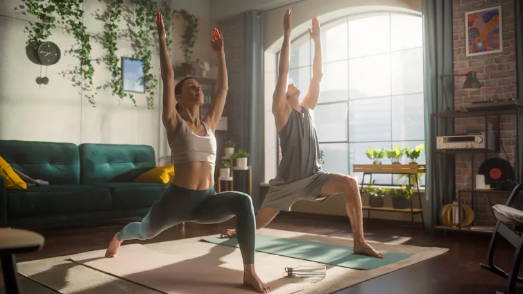 A fit man and woman perform a yoga move in an apartment while the morning sun shines through a window.