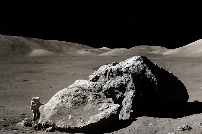Photo of the surface of the moon. An astronaut in a spacesuit stands next to a large rock. A moon buggy is parked on the other side of the rock.