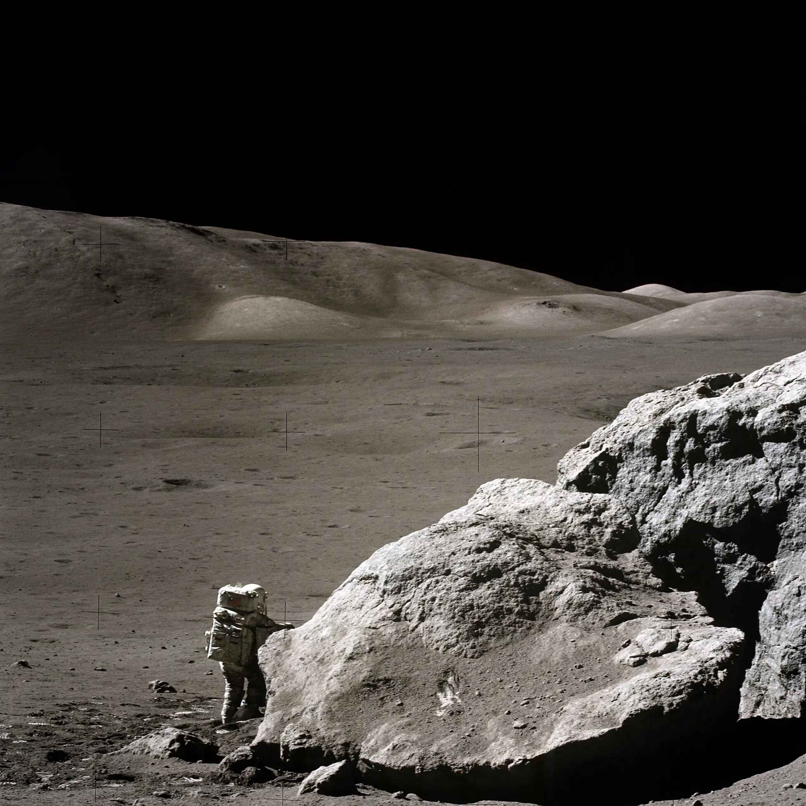 Photo of the surface of the moon. An astronaut in a spacesuit stands next to a large rock. A moon buggy is parked on the other side of the rock.