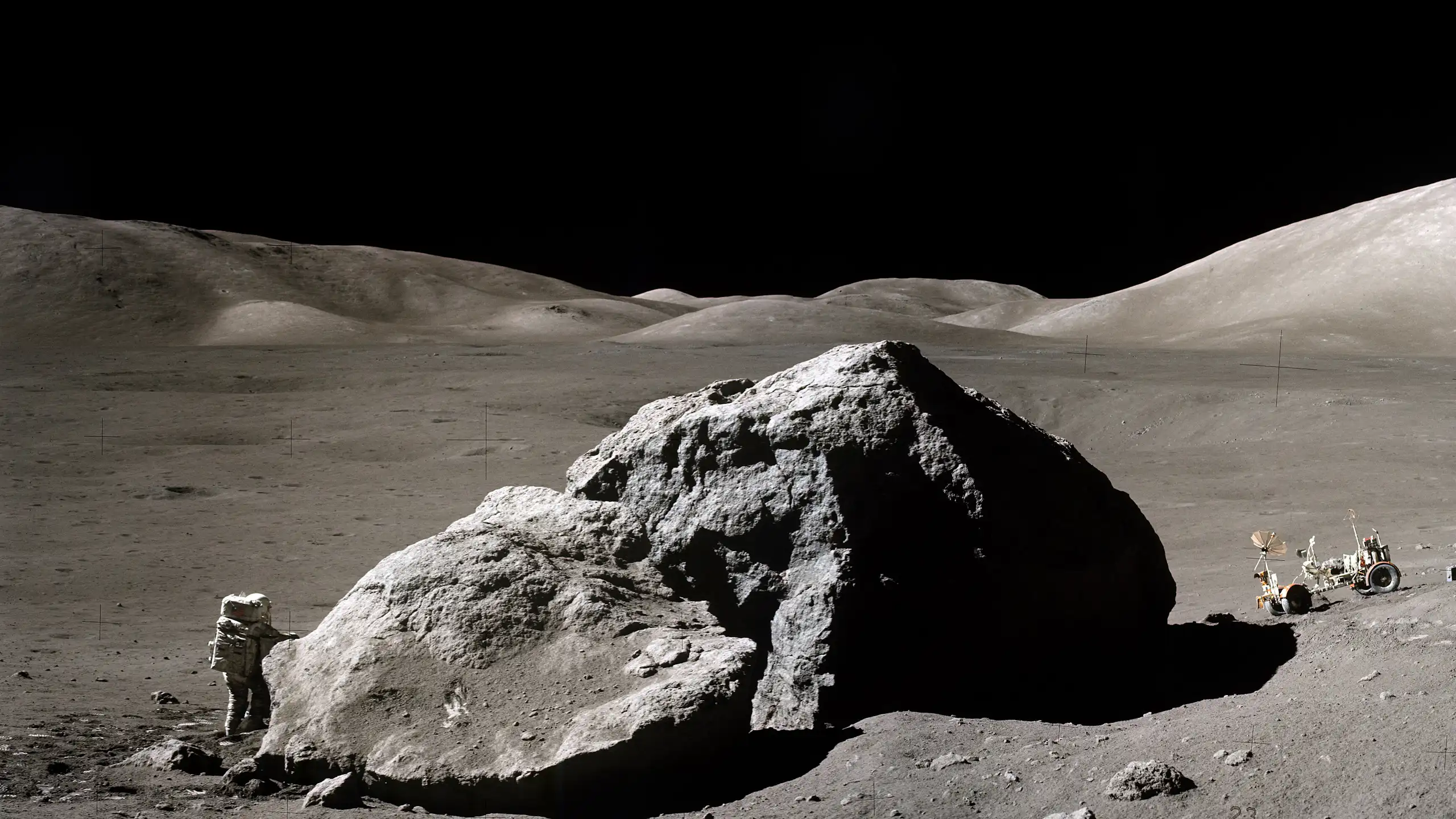 Photo of the surface of the moon. An astronaut in a spacesuit stands next to a large rock. A moon buggy is parked on the other side of the rock.