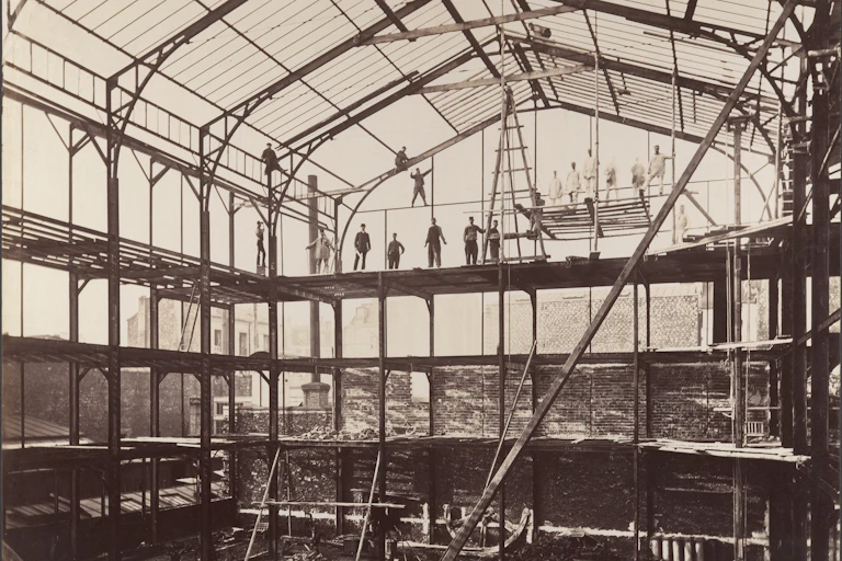 photo en noir et blanc d'un hangar en ver et métal avec des ouvriers en équilibre sur des poutres
