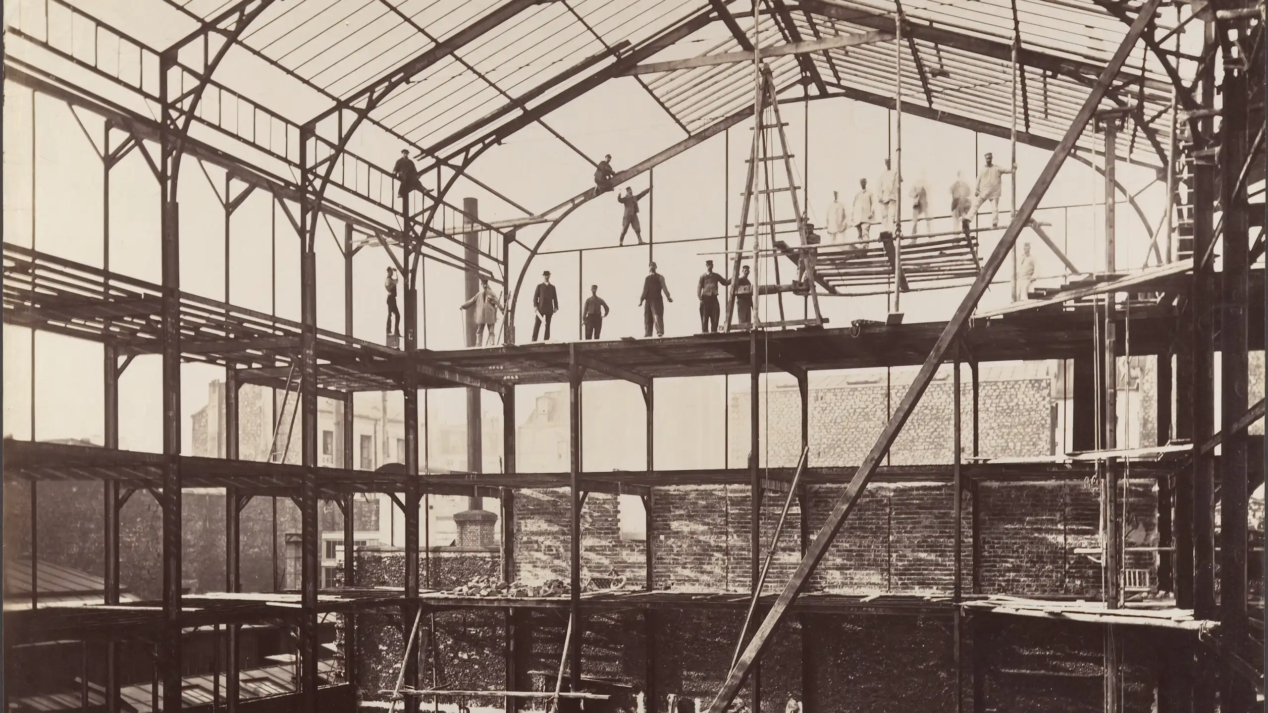 photo en noir et blanc d'un hangar en ver et métal avec des ouvriers en équilibre sur des poutres